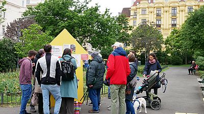 Menschen stehen in einem Park und betrachten eine gelbe Ausstellungstafel