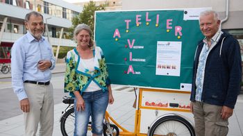 Zwei ältere Männer und eine Frau stehen vor einer Tafel auf einem Lastenrad und lächeln