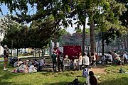 Eine Gruppe von Menschen sitzt im Schatten hoher Bäume auf einer Wiese und schaut einer Freiluft-Theateraufführung mit rotem Vorhang zu. Im Hintergrund sind ein Sportplatz und städtische Wohnhäuser zu sehen.