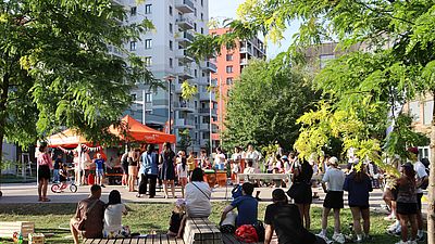 viele Menschen sitzen und stehen draußen bei Bäumen vor Gebäuden