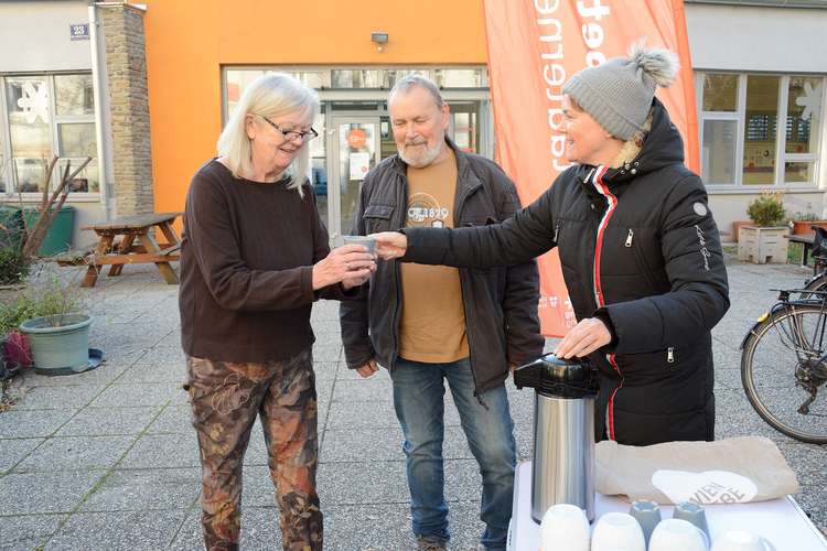 Eine GB*Mitarbeiterin überreicht eine Tasse an ein älteres Ehepaar.