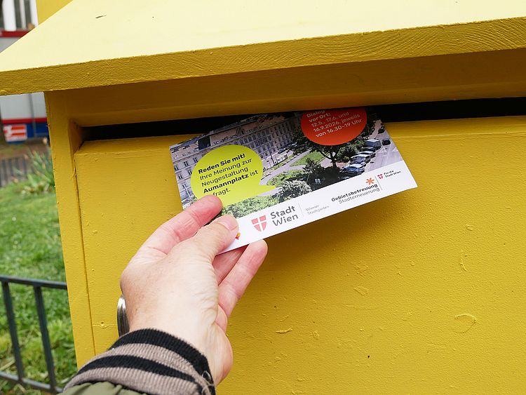 Eine Hand wirfst eine Postkarte in einen gelben Briefkasten.