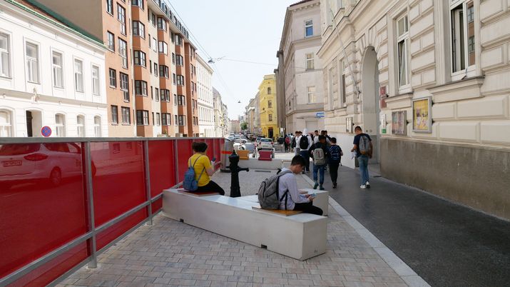 Neuer Schulvorplatz Leopold-Ernst-Gasse neu gepflasterter Platz mit grauen Sitzmöbel, auf dem SchülerInnen sitzen, links ein rotes Geländer, rechts ein beiges Schulgebäude aus der Gründerzeit