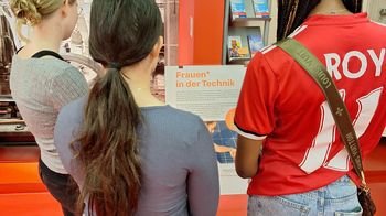 drei junge Frauen* lesen Plakat zu Frauen* in der Technik