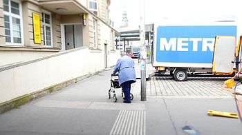 Person mit Rollator auf Gehsteig, dahinter steht ein LKW auf einem Parkplatz