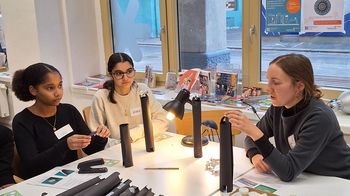 Drei Personen, weiblich gelesen, sitzen um Tisch mit Lampen und schwarzen Kegeln