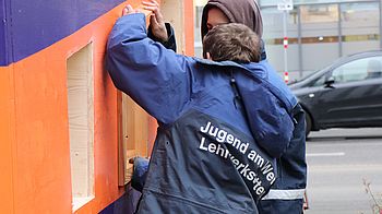 Zwei Personen von Jugend am Werk bauen Fenster in ein Stadtmöbel ein