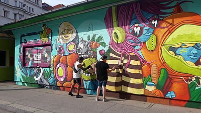 bunte Wand mit Graffiti, zwei Personen sprühen an die Wand