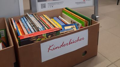 Eine Bückerkiste mit der Aufschrift "Kinderbücher" steht in einem Raum vor einem A-Ständer, indem ein Plakat zur Bewerbung des Büchertauschfreitags im GB*Stadtteilmanagements steht.