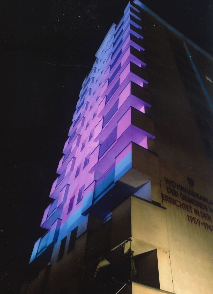 Das Bild zeigt ein hohes Wohnhaus bei Nacht, das mit farbigem Licht angestrahlt wird. Die Fassade ist in kräftigen Tönen von Lila und Blau beleuchtet, was dem Gebäude ein künstlerisches, fast surreal wirkendes Aussehen verleiht. Die Balkone werfen markante Schatten und betonen die Struktur des Hauses. An der rechten Seite des Gebäudes ist ein Text in deutscher Sprache zu sehen, darunter unter anderem „WOHNHAUSANLAGE DER GEMEINDE… ERRICHTET IN DEN JAHREN 1959–1961“. Der Himmel im Hintergrund ist dunkel, fast schwarz, was die Lichtinstallation besonders hervortreten lässt.
