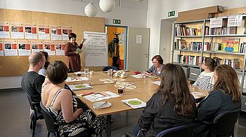 Menschen sitzen rund um einen Holztisch und schauen auf eine stehende Person, die gerade an einem Flipchart die eingereichten Ideen präsentiert.