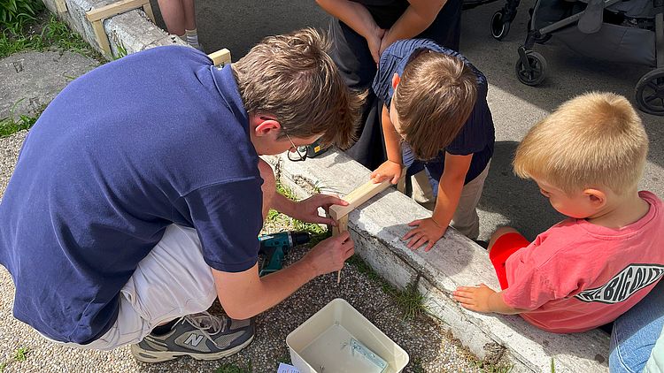 zwei Kinder und eine erwachsene Person, alle männlich gelesen und weiß, betrachten Holzstücke und messen aus