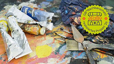 Man sieht Acryl-Farbtuben und Pinsel auf einem bunt gemusterten Tisch.