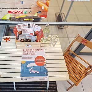 Holzklappstuhl rechts von einem weißen Holzklapptisch. Auf dem Tisch liegen in einer Kiste Infomatierialien zum Thema Hitze in der Stadt und zum Projekt. Auch ein Plakat, das auf den Sommer-Rastplatz aufmerksam macht, liegt auf dem Tisch.