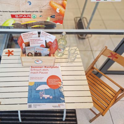 Holzklappstuhl rechts von einem weißen Holzklapptisch. Auf dem Tisch liegen in einer Kiste Infomatierialien zum Thema Hitze in der Stadt und zum Projekt. Auch ein Plakat, das auf den Sommer-Rastplatz aufmerksam macht, liegt auf dem Tisch.
