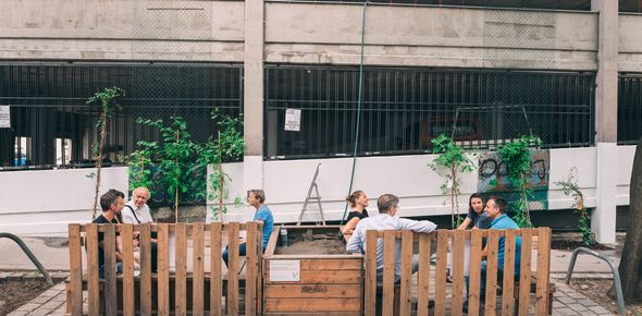 Menschen sitzen in einem Parklet aus Holz vor einer Hochgarage aus Beton