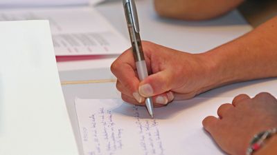 Bildausschnitt von beiden Händen eines Menschen, der gerade an einem Tisch auf einem Blatt Papier etwas mit einem Kugelschreiber schreibt.