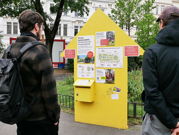 2 Personen stehen in einem Park und betrachten eine gelbe Ausstellungstafel