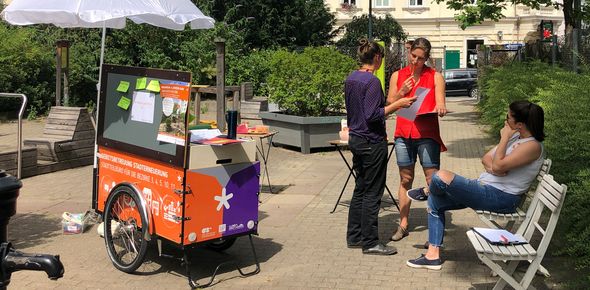 Lastenrad mit Infomaterial steht auf einem kleinen Platz im Park, daneben stehen zwei Personen, die miteinader sprechen, eine dritte sitzt daneben.