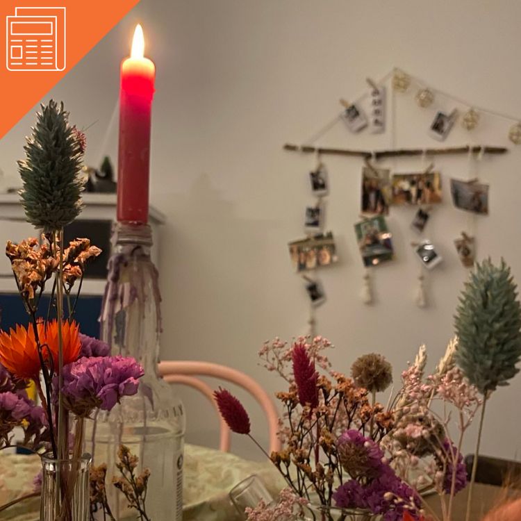 Ein rote brennden Kerze und Trockenblumen azf einem Tich, im Hintergund vescrhwommen ein Mobile mit Fotos