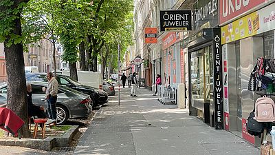 Foto von einem Gehweg im 10. Bezirk. auf der linken Seite befinden sich abwechselnd parkende Autos und Bäume. Rechts die Erdgeschosslokale in Gründerzeitbauten.