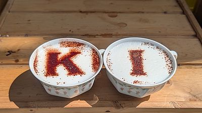 Zwei Kaffeetassen stehen auf einer Holzoberfläche. In den Milchschaum ist mit Kaokao jeweils ein Buchstabe gepudert: einmal K einmal I.
