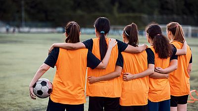 Junge Fußballer*innen stehen Arm in Arm vorm Fußballfeld. 