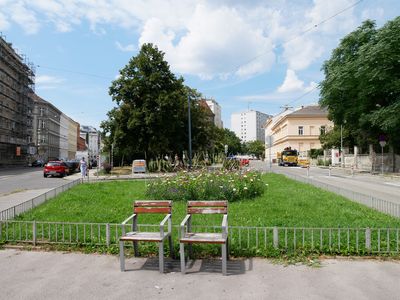 Mahrere Grünflächen werden zu einem Park Grünfläche umgeben von Straßen, davor zwei Stühle