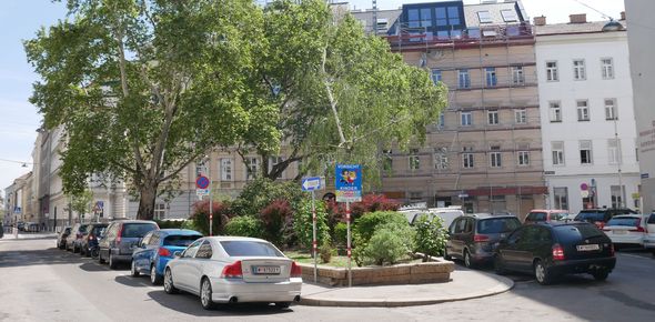 Kleiner urbaner Platz mit Bäumen, rundherum parkende Autos.