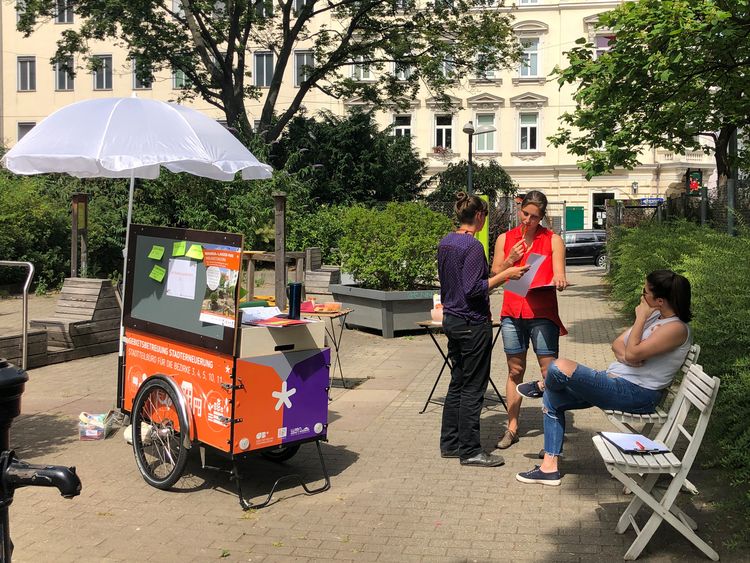 Lastenrad mit Infomaterial steht auf einem kleinen Platz im Park, daneben stehen zwei Personen, die miteinader sprechen, eine dritte sitzt daneben.