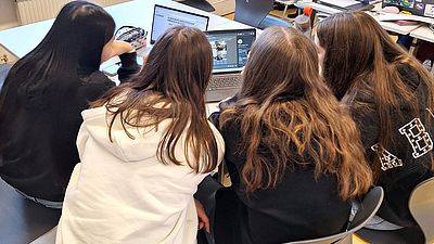 vier junge Personen, weiblich gelesen mit langen Haaren sitzen um Laptop und arbeiten