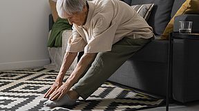 Seniorin, die sich auf einer Couch sitzend strecht und die Fingerspitzen zu ihren Zehen streckt.