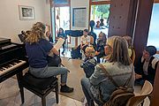 Musikunterricht in einer Musikschule: Eine Lehrerin spielt Gitarre vor einer Gruppe von Kindern und Erwachsenen. Im Vordergrund steht ein Flügel, im Hintergrund sind Streichinstrumente zu sehen.