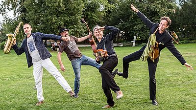 Vier Musikanten in witzigen, ausladenden Posen auf einer grünen Wiese. Jeder von den vier Männern hält ein Musikinstument in der Hand.