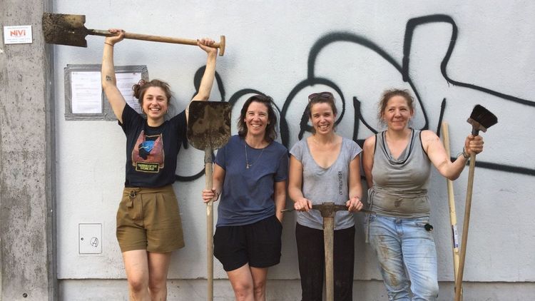 4 Frauen mit Schaufeln und Spaten stehen vor einer grauen Mauer