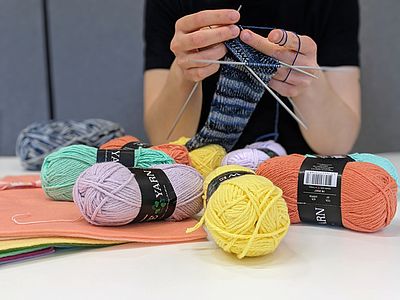 Bunter Garn auf einem weißen Tisch, im Hintergrund Strickt eine Frau.