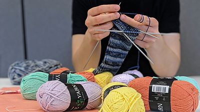 Bunter Garn auf einem weißen Tisch, im Hintergrund Strickt eine Frau.