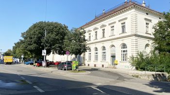 Weißes, altes Bahnhofsgebäude, daneben ein grüner Baum, davor Straße