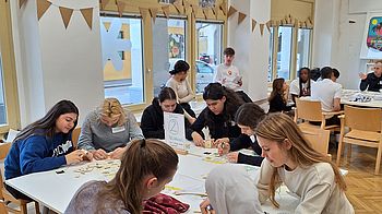 Großer Raum mit Tischen, an denen Menschen herumsitzen und etwas basteln. Girlanden im Hintergrund.