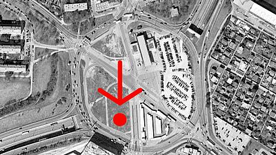 Schwarz-weißes Luftbild vom Verteilerkreis. Ein roter Pfeil und ein roter Punkt zeigen darauf, wo genau sich das Zwischennutzungsprojekt befindet.