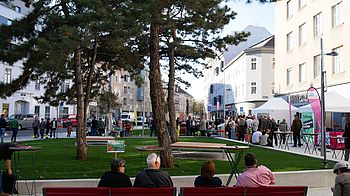 Der neue Dreiecksplatz Man sieht grünen Rasen, davor rote Sitzbänke auf denen Menschen sitzen, im Hintergrund Bäume