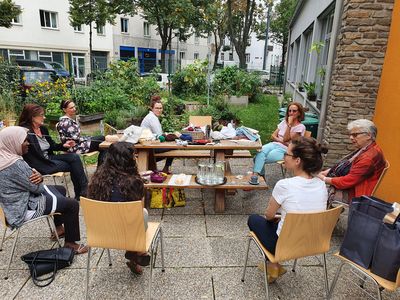 8 Frauen sitzen im freien rund um einen Tisch, im Hintergrund Hochbeete