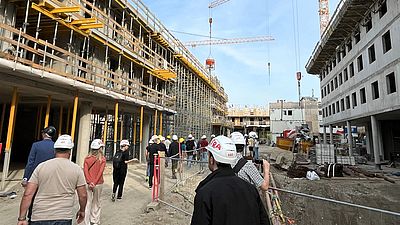 Menschen mit Baustellenhelmen sind auf einer Baustelle, im Hintergrund ist der Rohbau sichtbar.