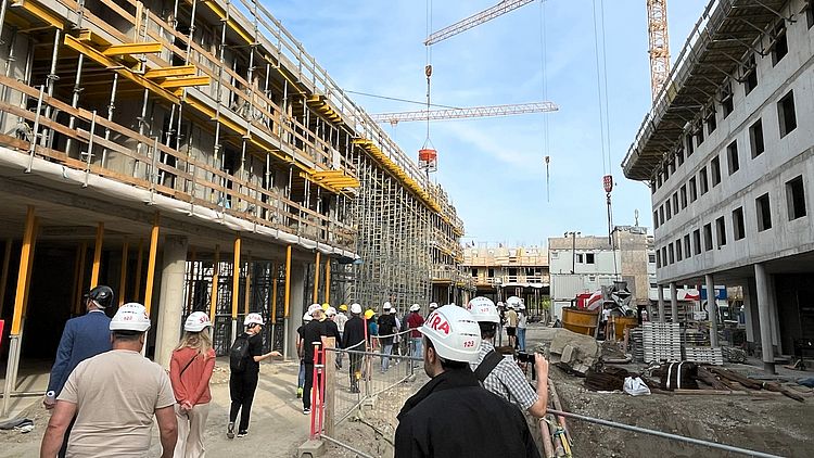 Menschen mit Baustellenhelmen sind auf einer Baustelle, im Hintergrund ist der Rohbau sichtbar.