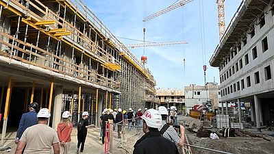 Menschen mit Baustellenhelmen sind auf einer Baustelle, im Hintergrund ist der Rohbau sichtbar.