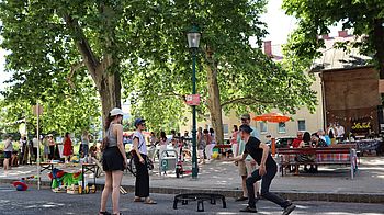 Parkkulisse mit mehreren Personen, vier spielen mit Ball und Netz (Spikeball), Bäume, Schatten