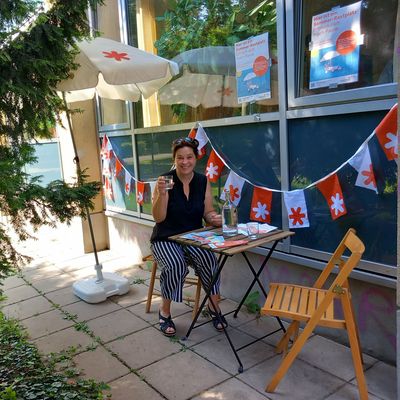 Eine Frau sitzt auf einem Holz-Klappstuhl. Neben ihr steht ein Holz-Klapptisch auf dem Infomatierialien zur Aktion "Sommer-Rastplätze" und zum Thema "Hitze in der Stadt" neben einer Wasserkaraffe und Trinkgläsern drapiert sind. Daneben ist ein weiterer, leerer Holzklappstuhl. Diese Szenerie befindet sich im Park neben der VHS Favoriten. An der Fensterfront der VHS sind zudem Wimpelketten, die mit dem GB*-Stern gebrandet sind, angebracht.