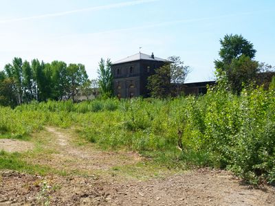 Freie Mitte Blick auf eine wilde , bewachsene Schotterfläche, im Hintergrund ein altes Backsteingebäude
