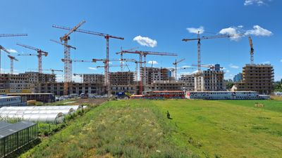 Hinter einer Wiese, links davon Glashäuser, befindet sich die Baustelle zum Quartier An der Schanze mit vielen Kränen und im Bau befindlichen Gebäuden.
