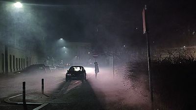 Fotografie von Michaela Hintermayr: Auto auf einer Art Landstraße in der Dunkelheit, Lichtkegel, die zeigen, wie nebelig es ist. Gegen das Licht ist auch der Umriss einer Person erkennbar.